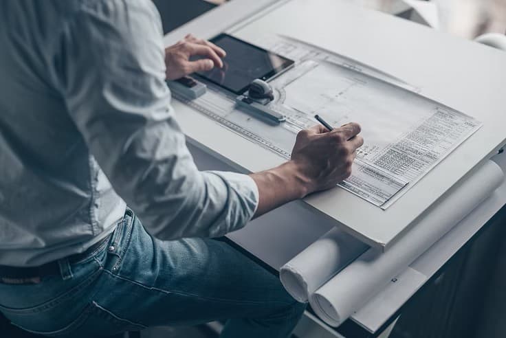Architect sketching on blueprints at a drafting table while simultaneously using a digital tablet.
